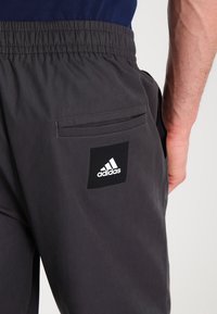 Mörkgrå träningsbyxor med elastisk midja, sidoficka och en svart rektangulär patch med en vit Adidas-logotyp.