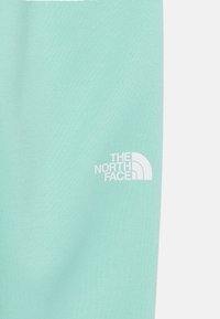 Svijetlo mint zelena tkanina s glatkom teksturom, koja sadrži bijeli logo "The North Face" u donjem lijevom kutu.