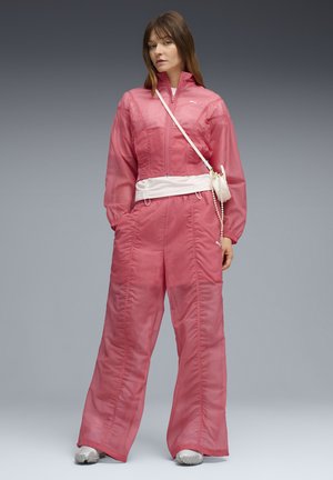 Woman wearing a pink translucent tracksuit with wide-leg pants, white sneakers, and a small white crossbody bag against a gray background.