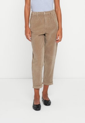 Kvinde iført beige kordfløjlsbukser, der er rullet op ved anklerne, kombineret med sorte balletsko og en lyseblå stribet top.