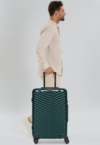 Man in beige outfit die loopt terwijl hij een donkergroene harde koffer met vier wielen en een uitklapbare handgreep trekt.