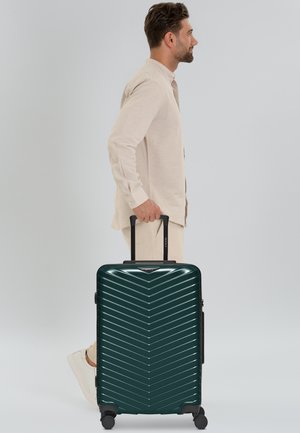 Uomo in abito beige che cammina mentre trascina una valigia rigida verde scuro con quattro ruote e manico estensibile.