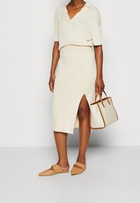 Une personne portant un haut en tricot couleur crème et une jupe midi assortie avec une fente sur le côté, tenant un sac beige avec des bordures marron, et des chaussures slip-on beiges.