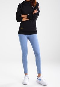 Kvinna står med armarna i kors, iförd svart huvtröja, ljusblå skinny jeans och vita Nike-sneakers med blå swoosh.