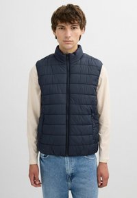 Chaleco acolchado azul marino con cuello alto, cierre de cremallera y patrón de acolchado horizontal. Lleva puesto sobre una camiseta de manga larga de color claro.