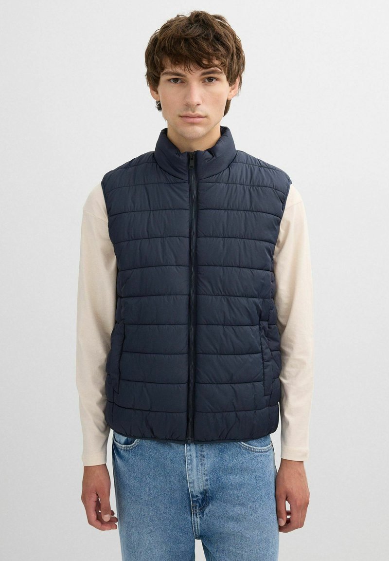 Chaleco acolchado azul marino con cuello alto, cierre de cremallera y patrón de acolchado horizontal. Lleva puesto sobre una camiseta de manga larga de color claro.