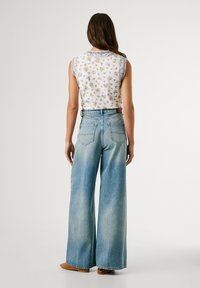 Bijela majica bez rukava s cvjetnim uzorkom u kombinaciji s svijetloplavim širokim trapericama. Traperice imaju visoki struk i stražnje džepove.