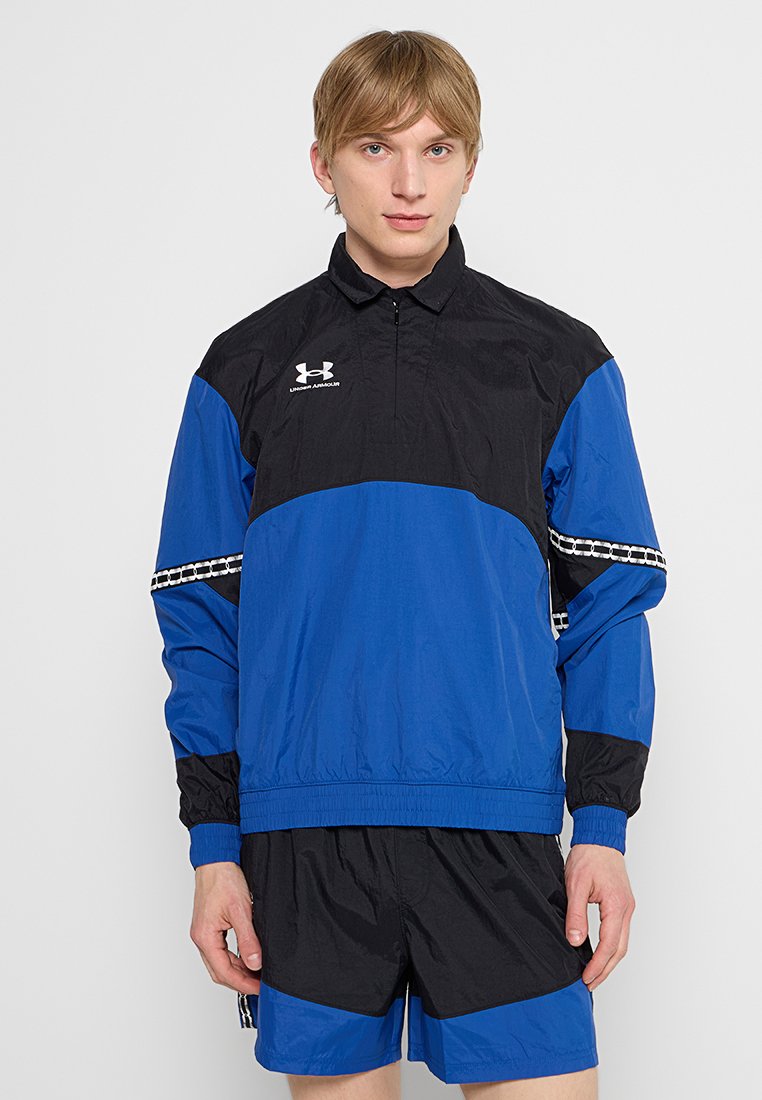 Chaqueta cortavientos con paneles negros y azules, cuello con cremallera, puños elásticos, logo de Under Armour y detalles a cuadros en las mangas.