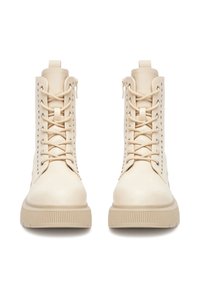 Beżowe buty wojskowe o gładkim wykończeniu, wysokiej cholewce, sznurowaniu z przodu i srebrnymi oczkami. Charakteryzują się grubą, teksturowaną podeszwą oraz tylną pętelką do zakładania.