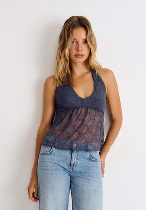 Junge Frau mit blondem Haar, die ein ärmelloses dunkles Spitzenoberteil und hellblaue Jeans trägt, steht vor einem weißen Hintergrund.