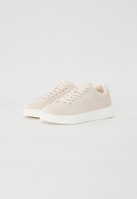 Lysebeige sueadesko med lav profil, flat hvit såle og matchende tåhette; har tradisjonell snøring.