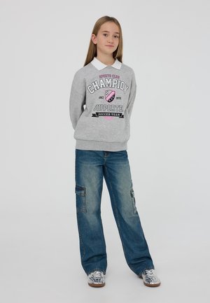 Felpa grigia con logo stampato "Champion Supporter", abbinata a jeans cargo larghi di colore blu chiaro con tasche. Sneakers con dettagli metallici.