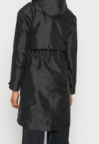 Veste noire imperméable avec taille cintrée, capuche et poignets boutonnés. Tissu lisse et brillant avec une coupe courte à l'arrière.