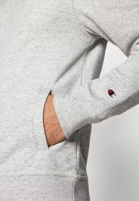 Grey sweatshirt with a soft texture, featuring a kangaroo pocket and a small embroidered logo on the sleeve. Simple, casual design.