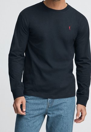 Mann trägt ein schwarzes Langarmshirt mit einem kleinen roten gestickten Logo auf der Brust und hellblauen Jeans.