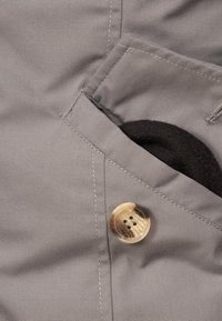 Šedá tkanina s hladkou texturou, s velkým knoflíkem a malou kapsou obsahující černý materiál. Švy jsou viditelné podél spojů.