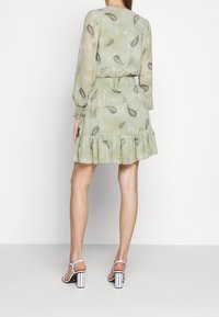 Grünes Kleid mit Paisleymuster, langen Puffärmeln und volantartigem Saum, aus leichtem Stoff, kombiniert mit weißen und schwarzen Absatzsandalen.