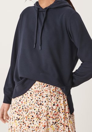 Kobieta ubrana w granatową bluzę z kapturem, zestawioną z dopasowaną do talii spódnicą o abstrakcyjnym, wielokolorowym wzorze.