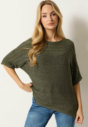 Jonge vrouw met lang blond haar, gekleed in een olijfgroen, gestructureerd gebreid topje met korte mouwen en een blauwe spijkerbroek, staand tegen een effen lichte achtergrond.