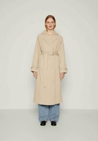 Trench coat bege com cintura cintuada, colarinho elegante e mangas longas. Apresenta um corte reto e detalhes mínimos, usado sobre jeans de perna larga.