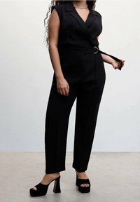 Black tailored jumpsuit with a fitted waist, v-neck, shoulder cutouts, and a belt. Paired with chunky black platform heels.