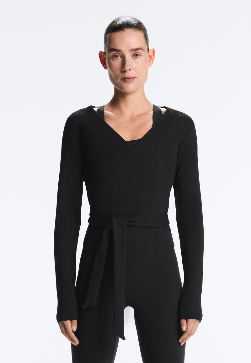 Femeie cu părul dat pe spate, purtând un pulover negru înfășurat, mulat, cu o centură legată, peste colanți negri, stând în picioare pe un fundal simplu.