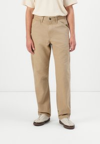 Beige cotton trousers with a straight-leg design, featuring two front pockets and a button closure. Worn with light-colored sneakers.