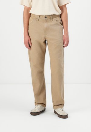 Pantalon en coton beige avec une coupe droite, doté de deux poches avant et d'une fermeture à bouton. Porté avec des baskets de couleur claire.