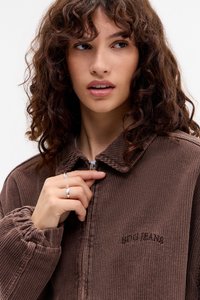 Joven mujer con cabello rizado castaño desabrochando una chaqueta de borra marrón con "BDG JEANS" bordado en el pecho, llevando anillos de plata en los dedos.