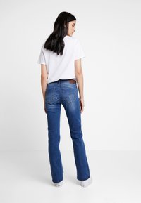 Kvinne med langt mørkt hår iført en hvit t-skjorte, blå jeans og hvite joggesko, stående med ryggen til mot en ensfarget hvit bakgrunn.