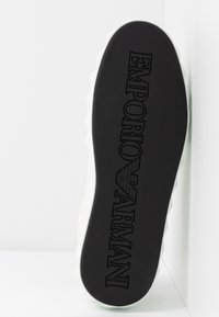 Musta kumisaapas, jossa on teksturoitu pinta ja kohotuilla kirjaimilla "EMPORIO ARMANI" -logo; sirot valkoiset reunat luovat kontrastia.