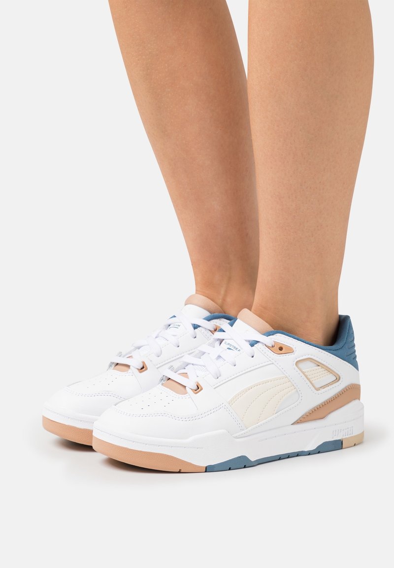 Baskets blanches avec des accents beige et bleu portées aux jambes inférieures sur un fond clair uni, montrant les vues de côté et de dessus.
