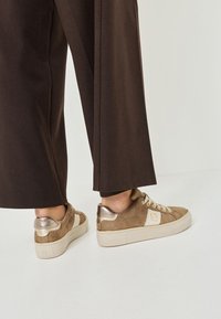 Tan mockasiner i mockaimitation med metallic guld accenter, texturerad detalj på hälen, och krämfärgade snören. Matchas med vida bruna byxor.