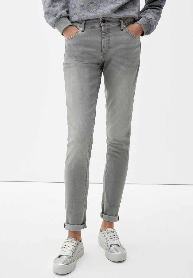 Vaqueros slim fit de color gris claro con bolsillos frontales y puños enrollados. Combinados con zapatillas bajas plateadas con cierre de cordones y acentos metálicos.