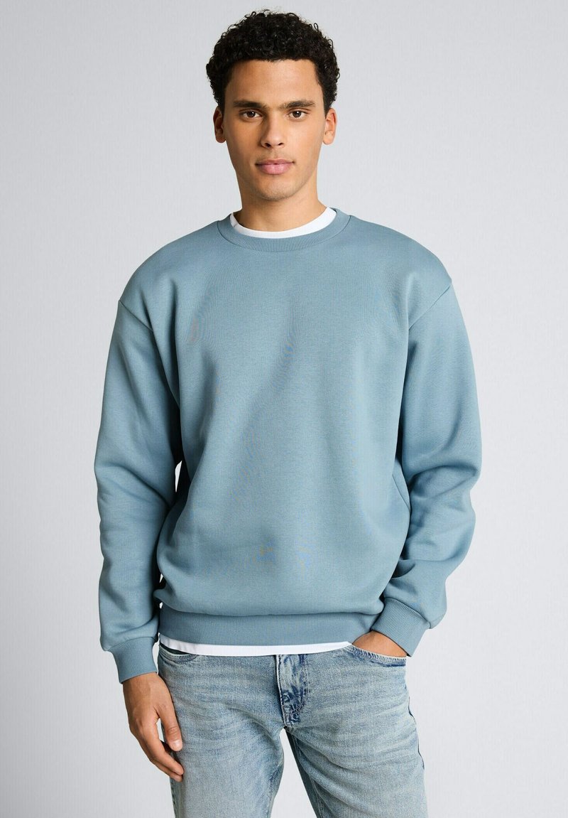 Junger Mann steht mit einer Hand in der Tasche, trägt ein hellblaues Sweatshirt und hellblaue Jeans vor einem einfarbigen Hintergrund.