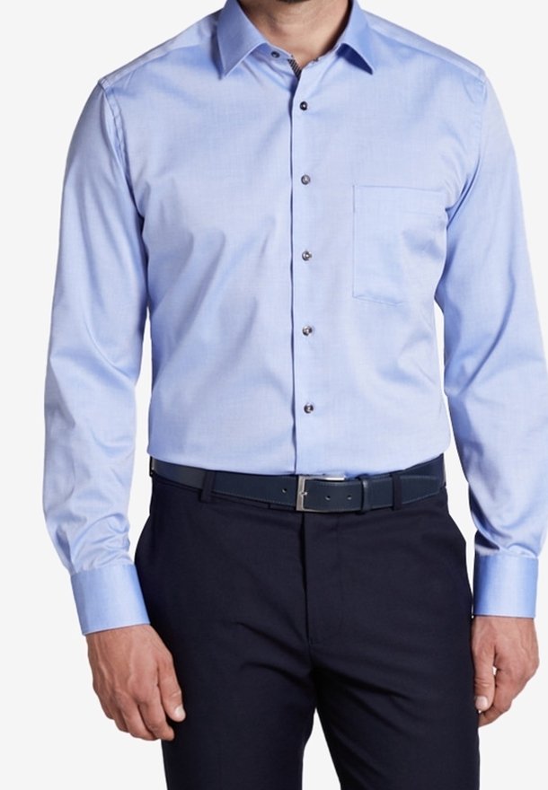 Homme portant une chemise bleu clair à manches longues avec une poche poitrine, rentrée dans un pantalon noir avec une ceinture noire.