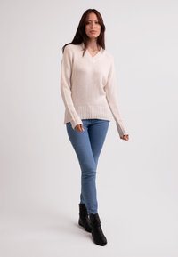 Beiger V-Ausschnitt-Pullover mit geripptem Saum und Bündchen, kombiniert mit hellblauen Skinny-Jeans und schwarzen Stiefeletten. Glatte Strickstruktur.
