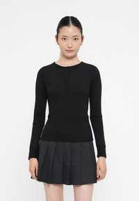 Jonge vrouw met donker haar vastgemaakt draagt een zwarte blouse met lange mouwen en een donkergrijze plissérok, staand tegen een witte achtergrond.