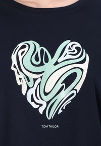 Tmavě modré bavlněné tričko s bílým a světlým zeleným srdcovým grafickým motivem a plynulými, abstraktními vzory; obsahuje logo 'TOM TAILOR'.