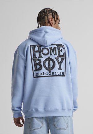 Persona con cabello trenzado que lleva una sudadera con capucha azul claro con el texto "HOME BOY loud•couture" en la espalda, combinada con vaqueros de mezclilla claros.