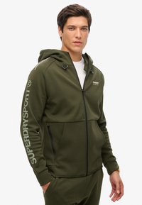 Felpa con zip color verde oliva, dotata di tasche, cerniere nere a contrasto e logo bianco "SUPERDRY SPORT" sulla manica, con cappuccio regolabile con cordino.