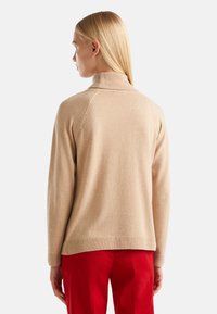 Femme aux cheveux blonds raides portant un pull beige à col roulé et un pantalon rouge, vue de dos sur un fond uni.