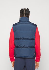 Granatowa pikowana kamizelka z wysokim kołnierzem, pikowanym wzorem i naszywką z logo u dołu, noszona na czerwonej koszuli z długim rękawem. Gładka faktura.