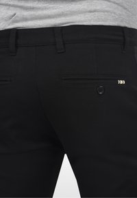 Zwarte broek met een gladde textuur, voorzien van een achterzak met knoopsluiting en gestreepte accenten. Lichtgewicht stof voor comfort.