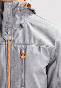 Grijze jas met een hoge kraag, oranje ritsaccenten, gestructureerde stof en een borstzak met een feloranje treklipje.