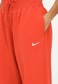 La mano derecha en el bolsillo de unos pantalones de chándal Nike de un rojo brillante, con cintura ajustable y logo blanco en la pierna, contra un fondo blanco.