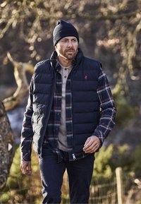 Mornarska puhovka brez rokavov preko karo srajce, s kremno osnovo in temno rdečimi poudarki. Temno modre kavbojke in pletena kapa zaokrožita oblačilo.