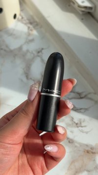 Main tenant un tube de rouge à lèvres MAC noir avec de longs ongles roses décorés sur une surface en marbre blanc.