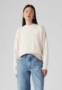 Felpa crema con scollatura rotonda e maniche lunghe, abbinata a jeans in denim azzurro chiaro a vita alta. Presenta un logo discreto sul petto.