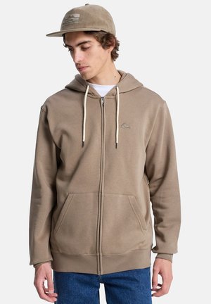 Brauner Reißverschluss-Hoodie mit einer Kapuze mit Kordelzug, zwei Fronttaschen und einem dezenten Logo. Kombiniert mit einer hellbraunen Mütze mit gestreiftem Design.